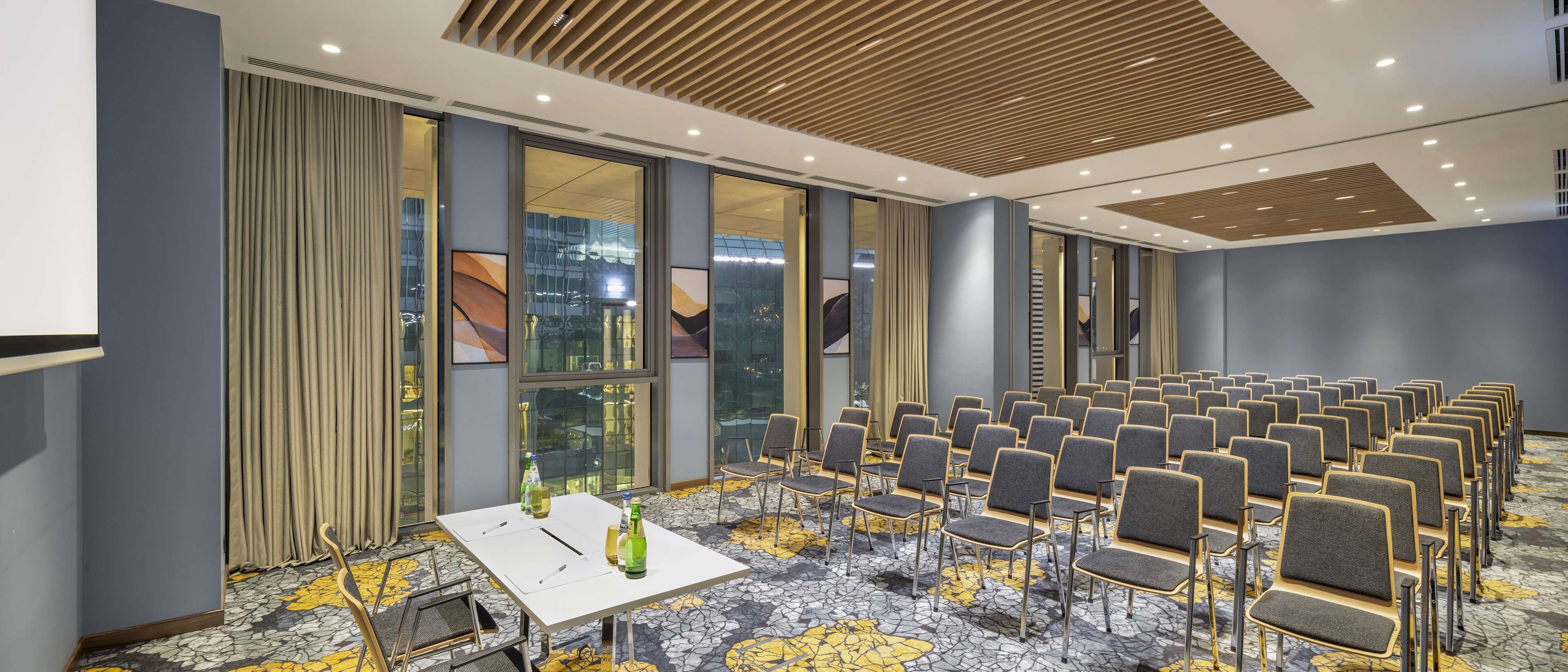 A conference room with rows of chairs, a table with bottles, and large windows with curtains.