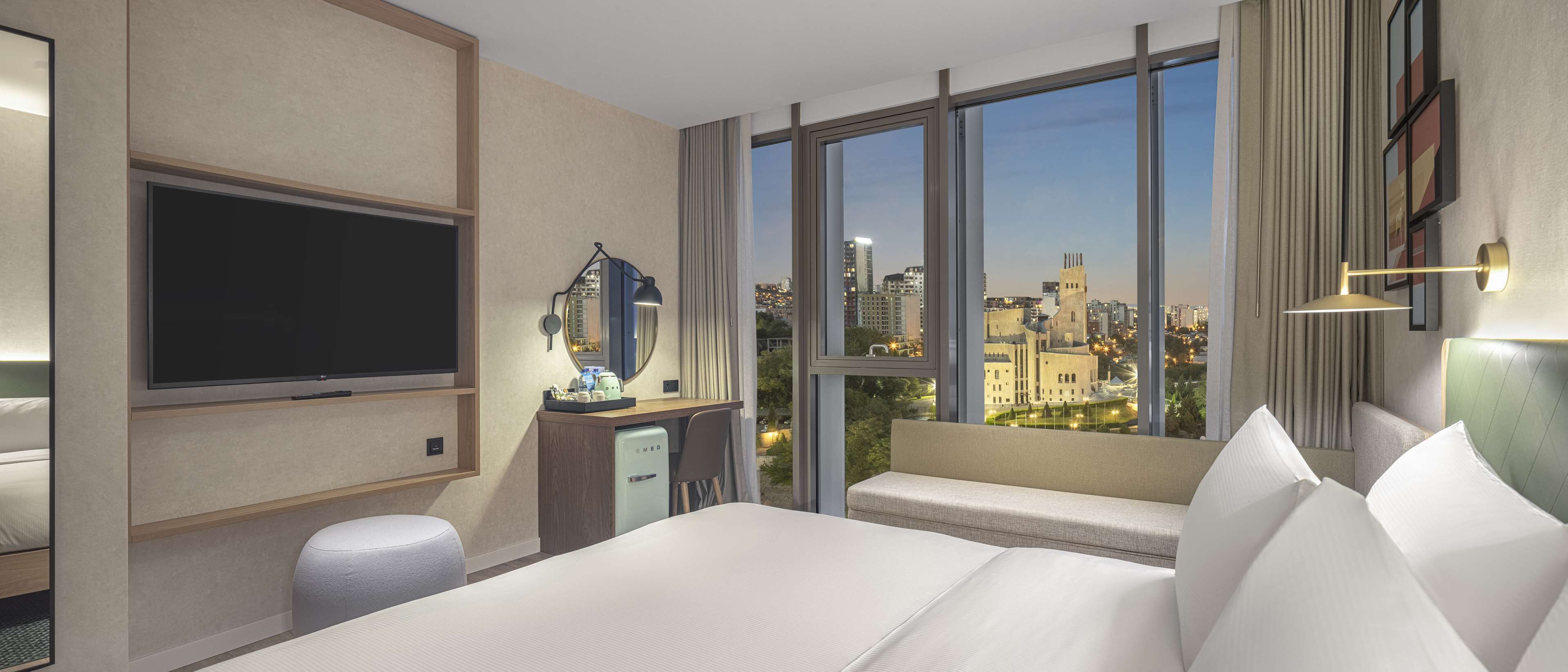 A hotel room with a large bed, a television, a desk with a lamp, a view of the cityscape through large windows, and a seating area by the window.