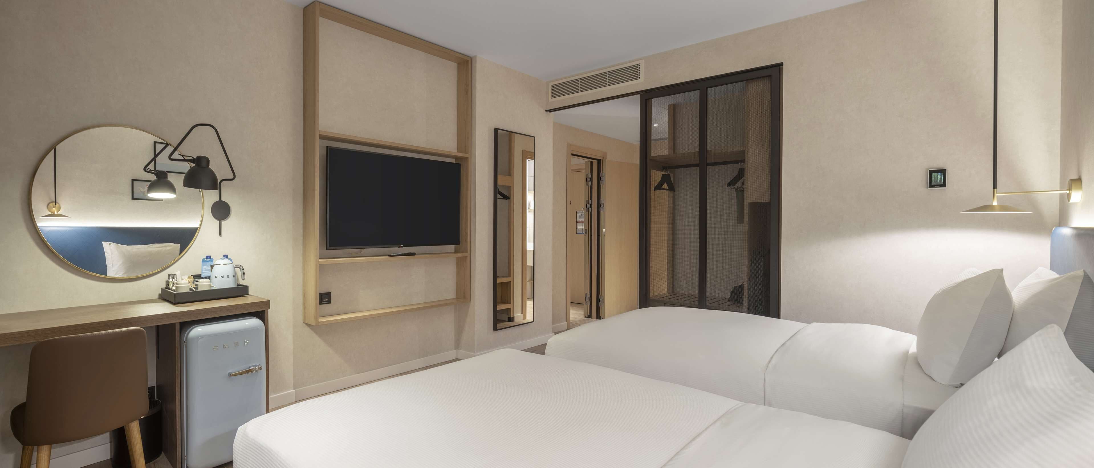 A hotel room with a bed, a desk with a computer, a mirror, a television, and a wardrobe.