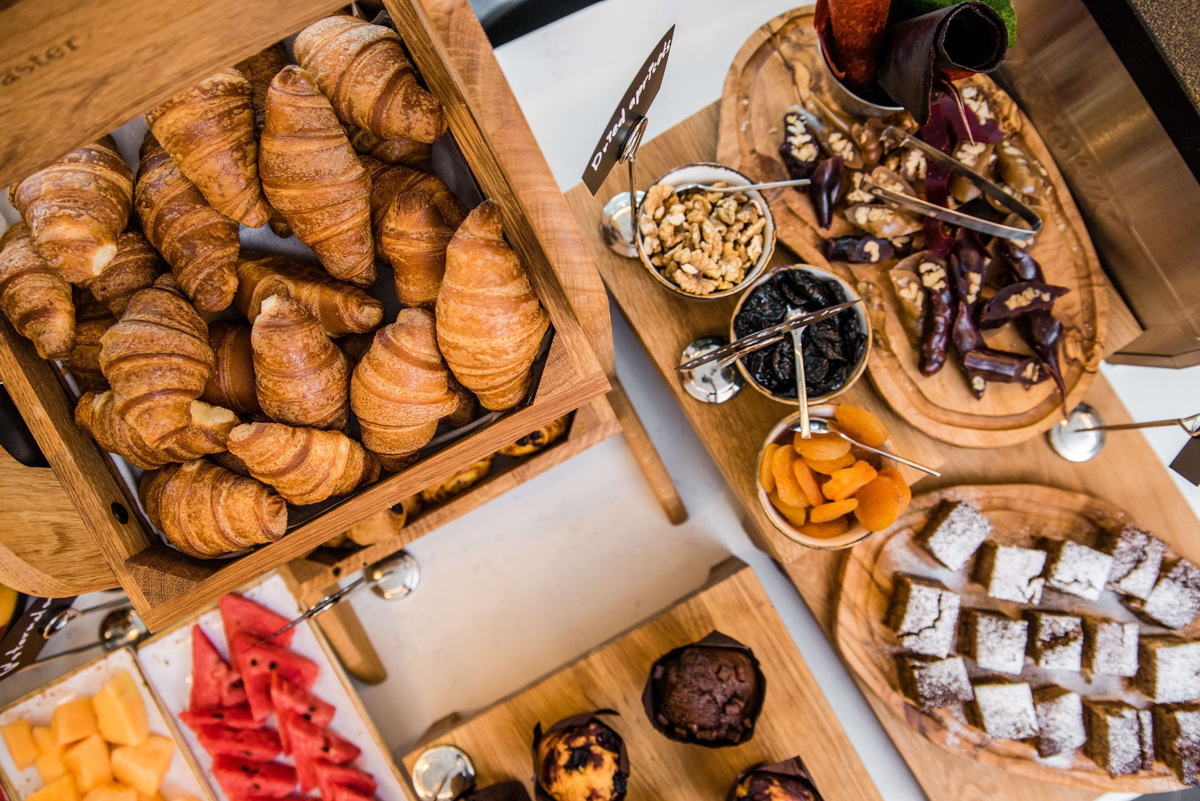 Diversos doces e sobremesas em bandejas de madeira, incluindo croissants, muffins e um bolo com açúcar em pó.