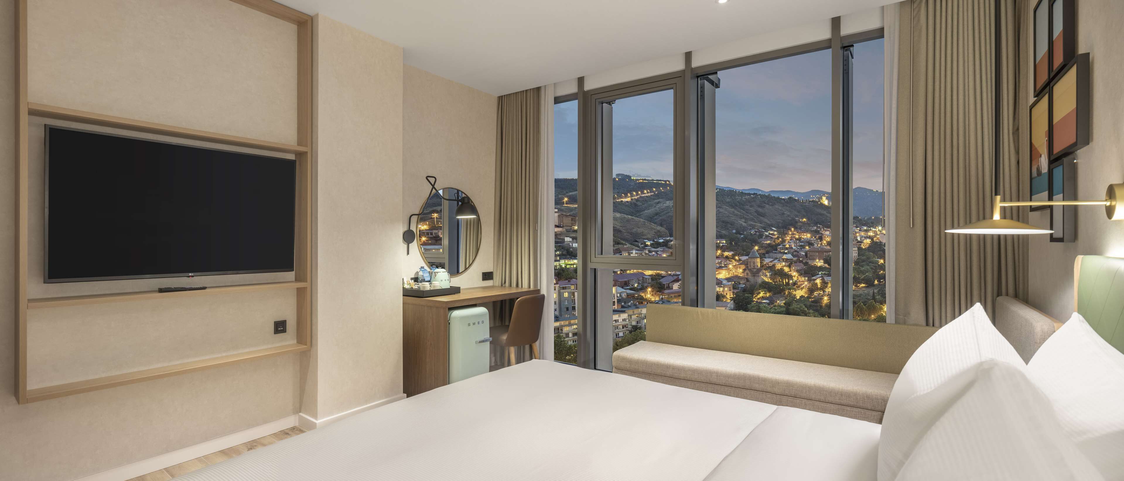 A hotel room with a bed, a TV, a desk with a mirror, and a view of a cityscape through large windows.