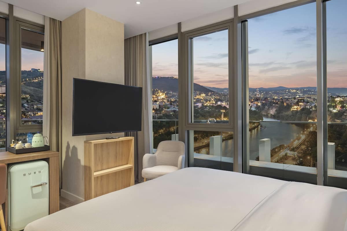 Een hotelkamer met een groot bed, een bureau met een computer, een televisie en uitzicht op een stadsbeeld.