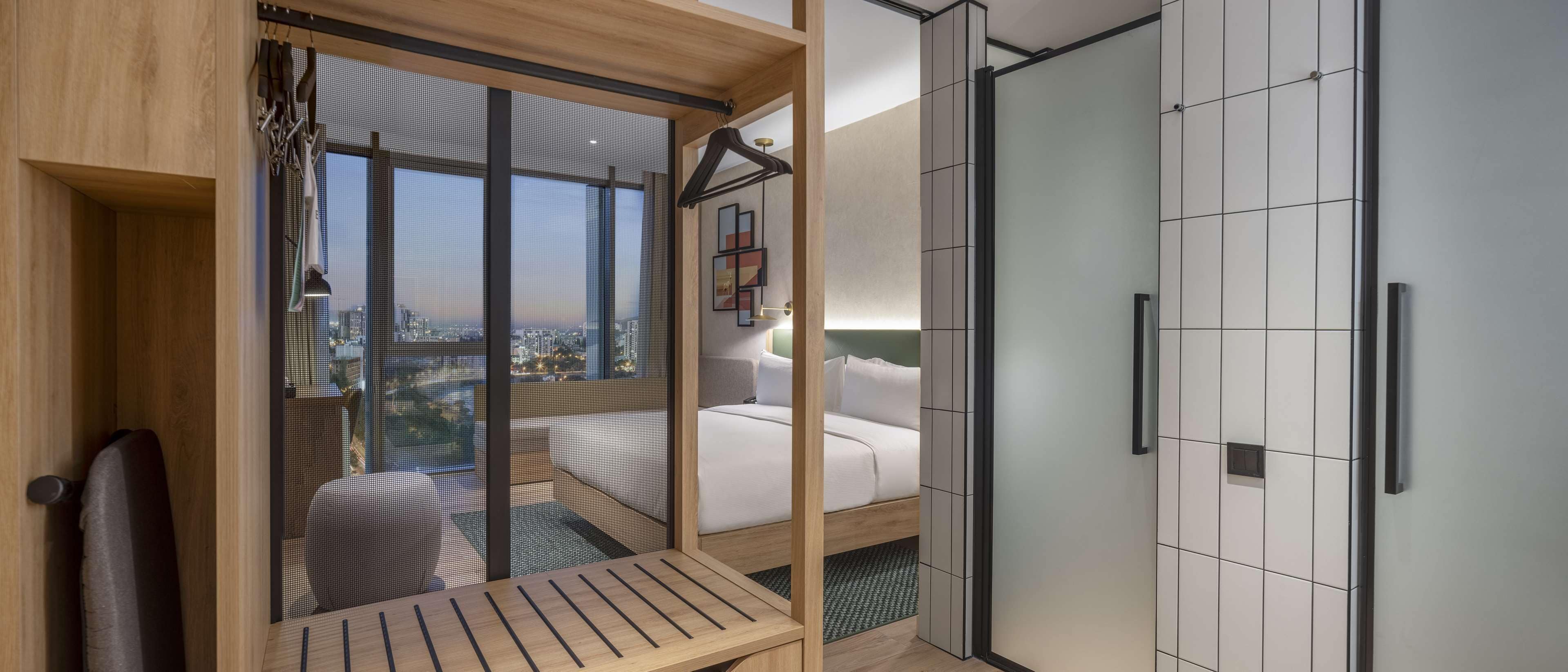 A modern bedroom with a city view, a bed, a bench, and a walk-in closet.