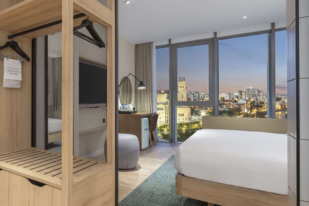 Een moderne hotelkamer met een groot bed, een houten garderobe, een zithoek met uitzicht, en een stadszicht door grote ramen.