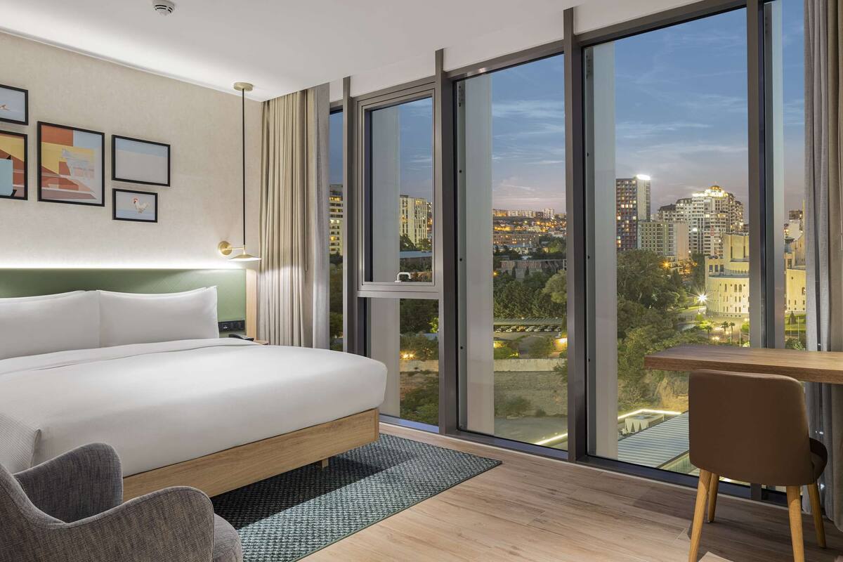 Een moderne hotelkamer met een groot bed, een bureau, een stoel en uitzicht op de skyline door middel van raam-tot-plafond ramen.