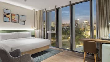 Een moderne hotelkamer met een groot bed, een bureau, een stoel en uitzicht op de skyline door middel van raam-tot-plafond ramen.