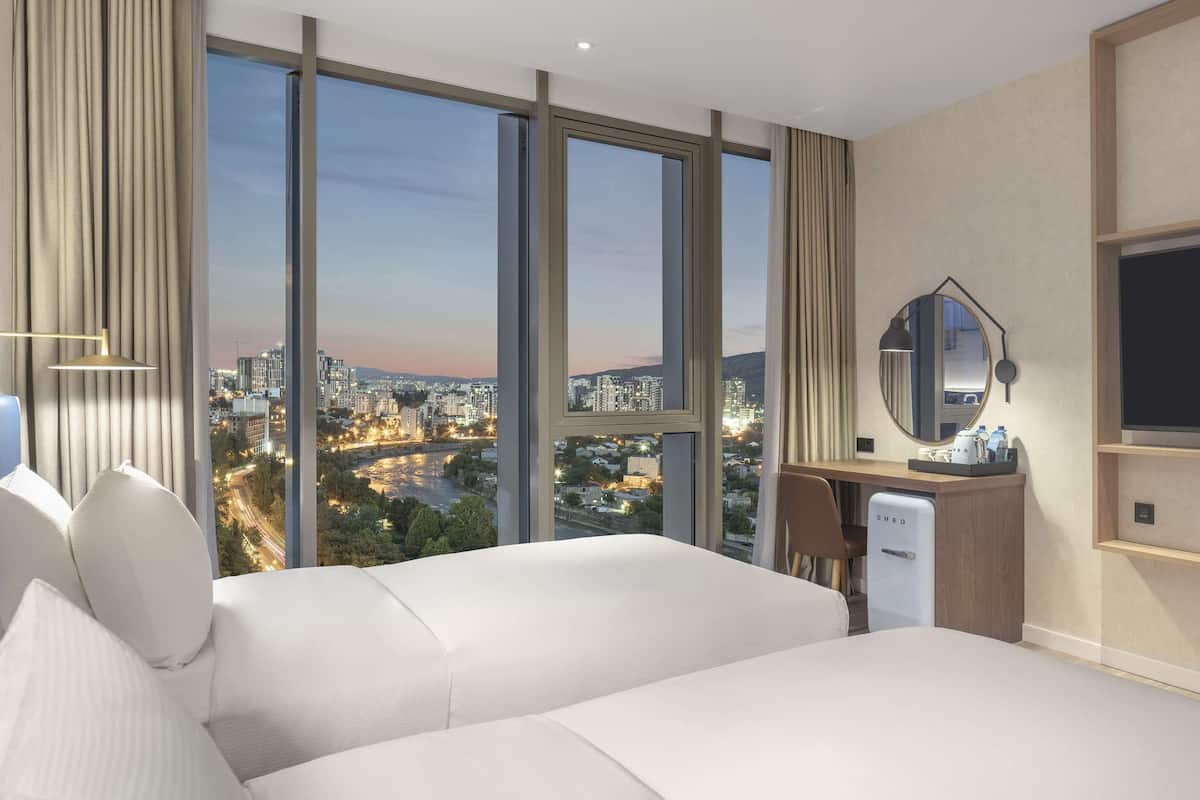Hotelkamer met een groot raam en uitzicht op de stad, een bed, een bureau met een koelkast en een wandgemonteerde flatscreen-tv.