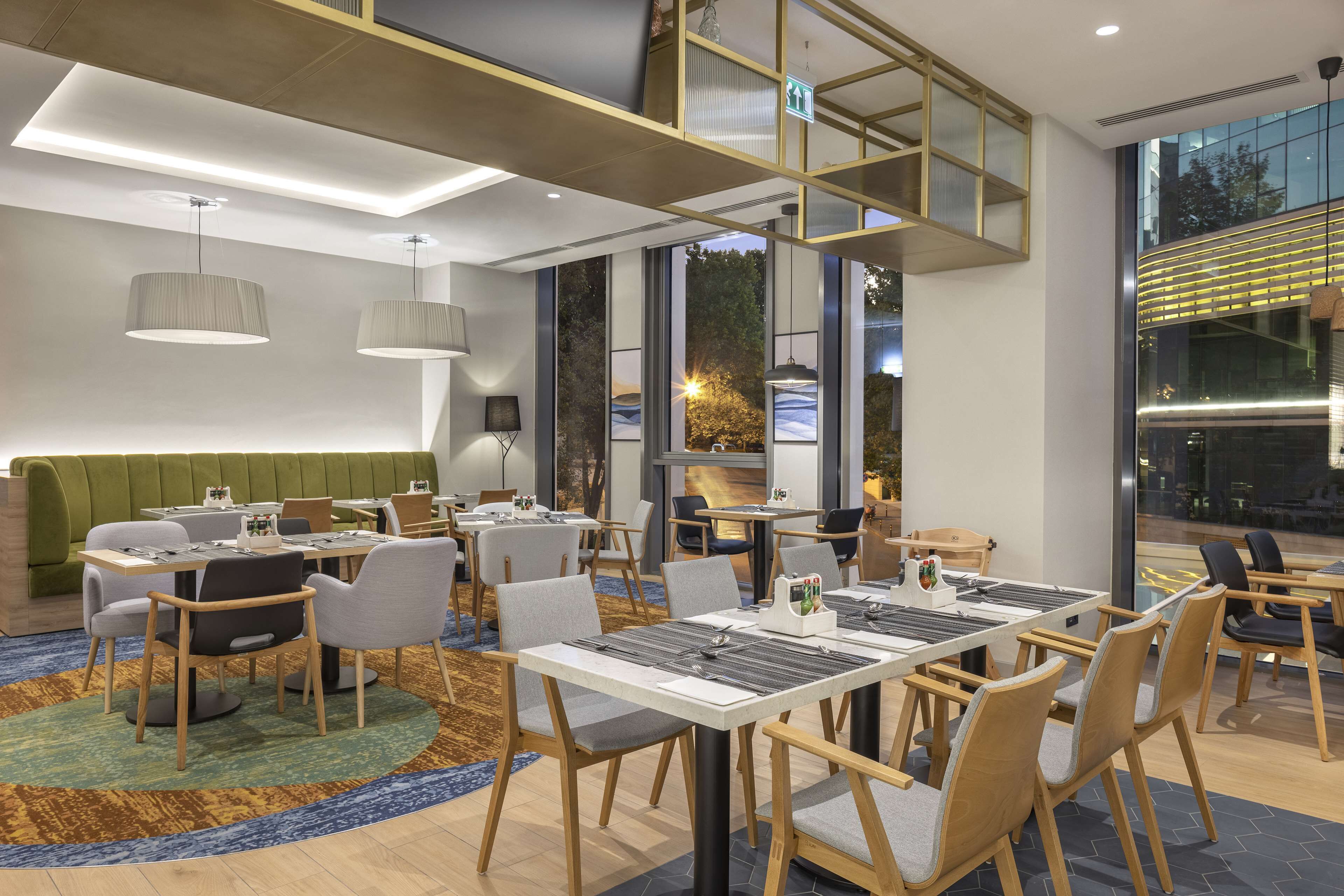 A modern dining area with multiple tables and chairs, a green cushioned bench, and large windows offering a view of the outdoors.