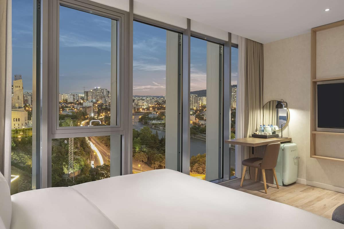 Een moderne hotelkamer met een groot bed, een bureau met stoel, een flatscreen televisie en stadszicht door grote ramen.