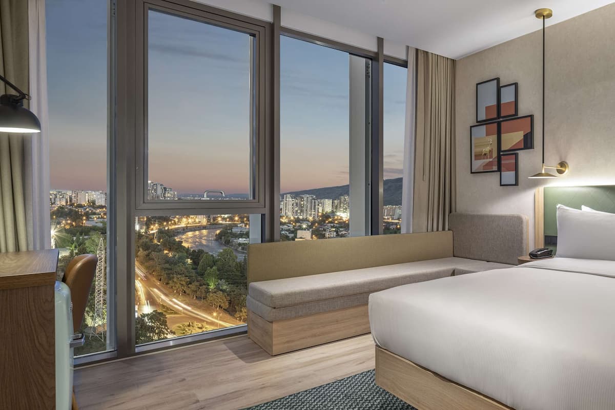 Een moderne hotelkamer met een groot bed, een zithoek en uitzicht op de stad door middel van kamerhoge ramen.