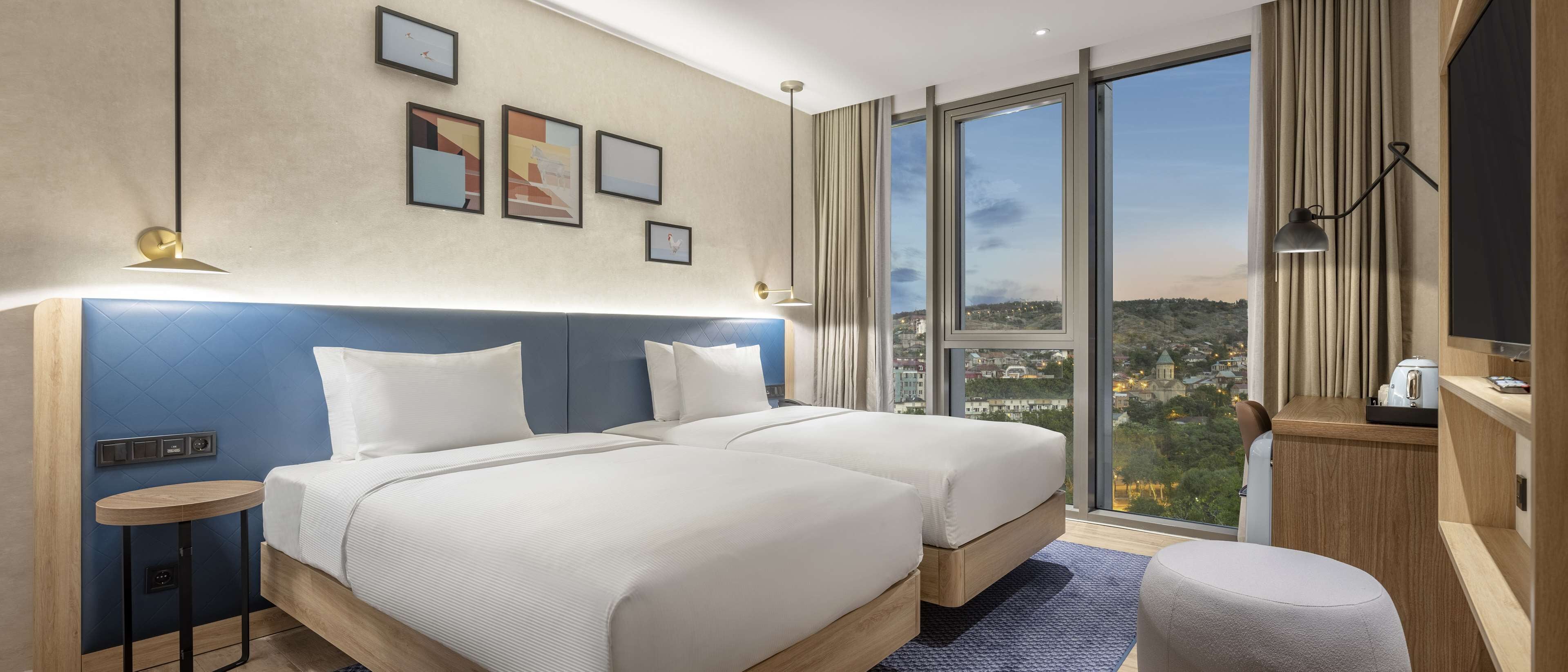 A hotel room with two beds, a large window with a city view, a desk with a lamp, and a TV.