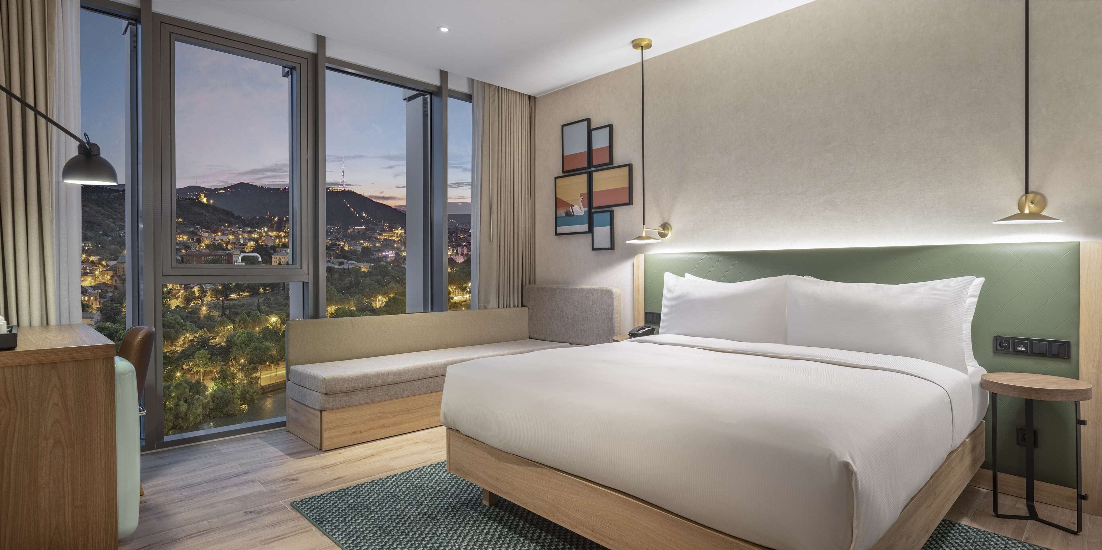 A modern hotel room with a large bed, a bench, and a view of a cityscape through large windows.