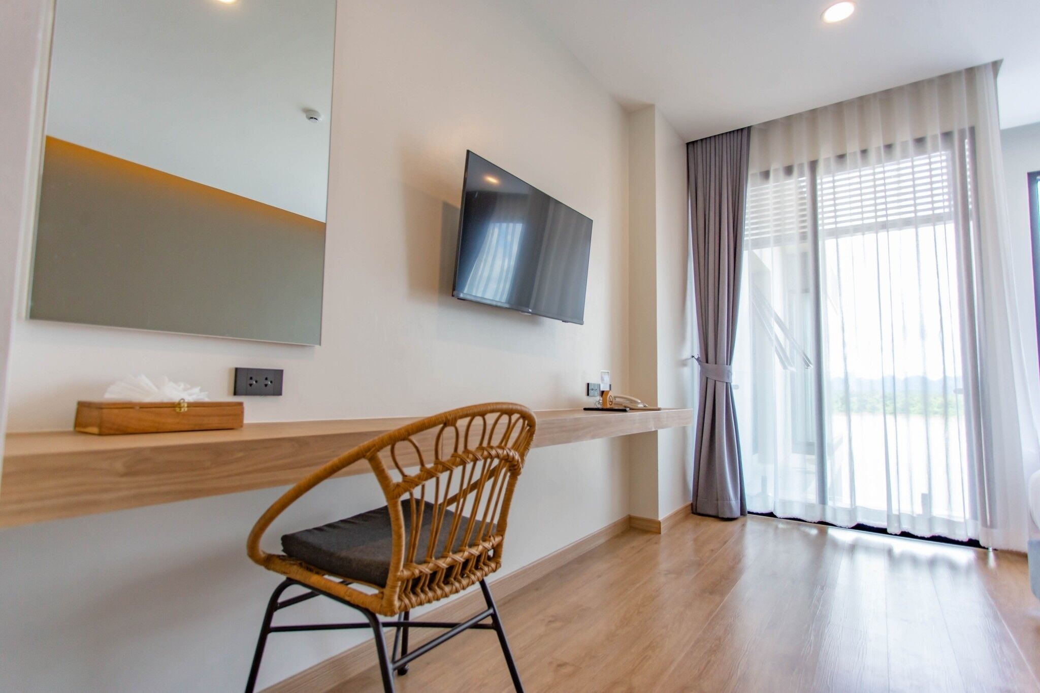 Ein modernes Hotelzimmer mit einem Holztisch, einem großen Spiegel, einem flachbildfernseher an der Wand und einem großen Fenster mit durchsichtigen Vorhängen.