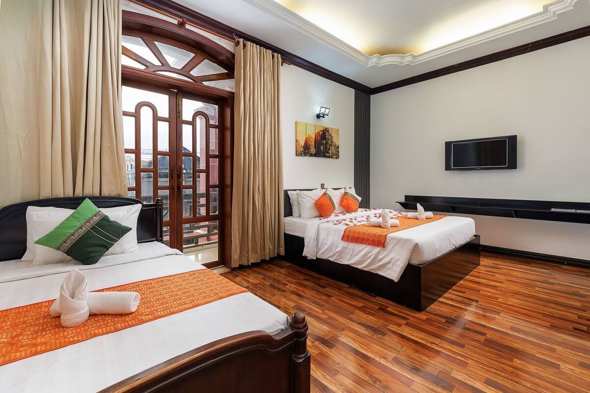A hotel room with two beds, wooden flooring, a TV, and a view through a glass door.
