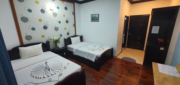 A hotel room with two single beds, a wooden floor, a wall with a clock pattern, a small bedside table with a vase, and a door with a sign.