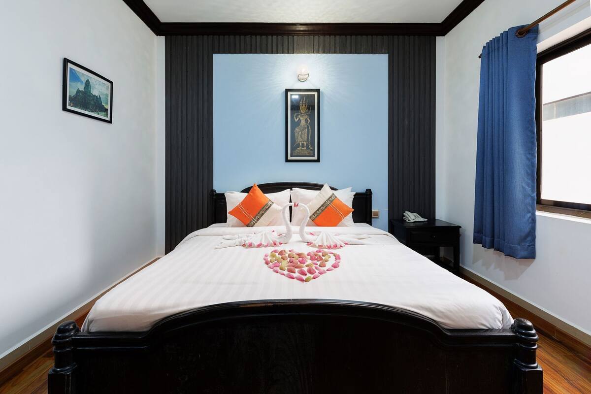 A neatly made bed with a heart-shaped arrangement of flowers, a framed picture on the wall, and a window with blue curtains.