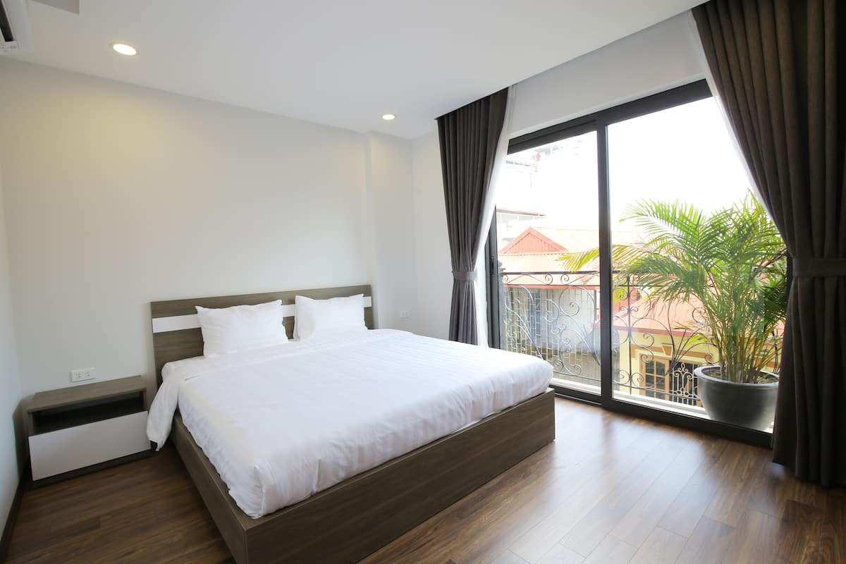 A modern bedroom with a large bed, a nightstand, a potted plant, and a view of the outside through a large window.