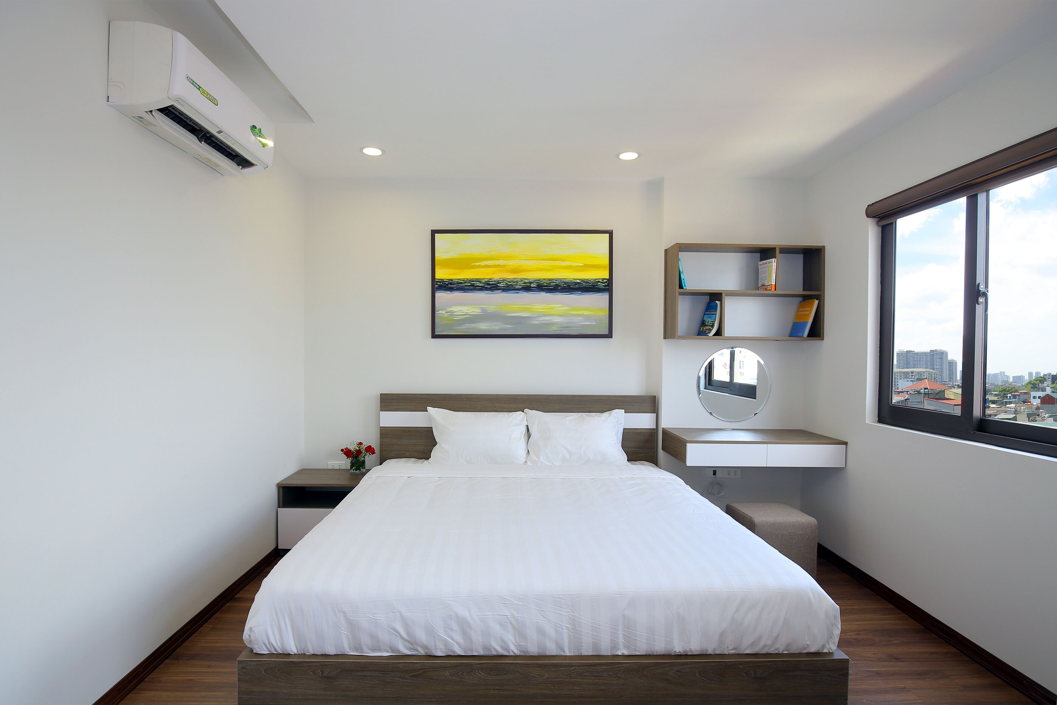 A modern hotel room with a large bed, a bedside table with a lamp, a small seating area, a bookshelf, and a window with a city view.