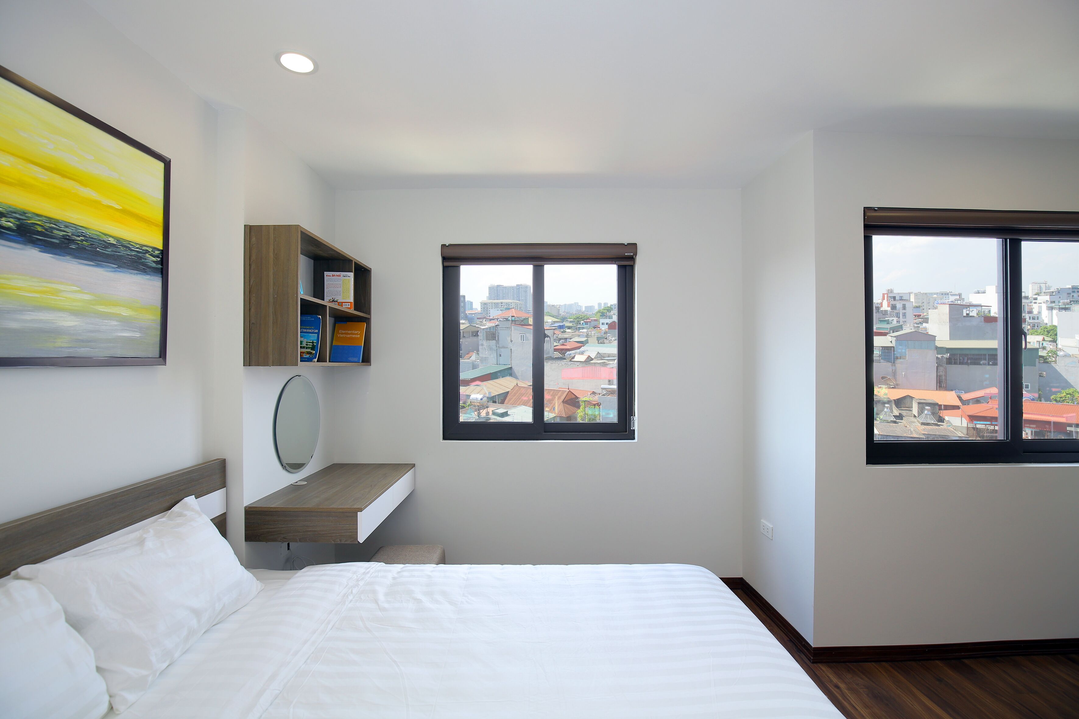 A hotel room with a bed, a small desk, a mirror, a bookshelf, and a window with a city view.
