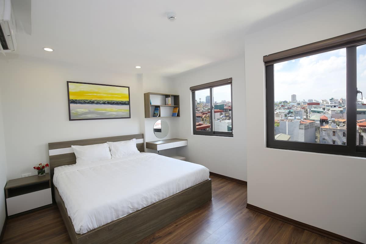 A modern bedroom with a large bed, a bedside table, a nightstand, a bedside mirror, a small shelf, and a window with a city view.