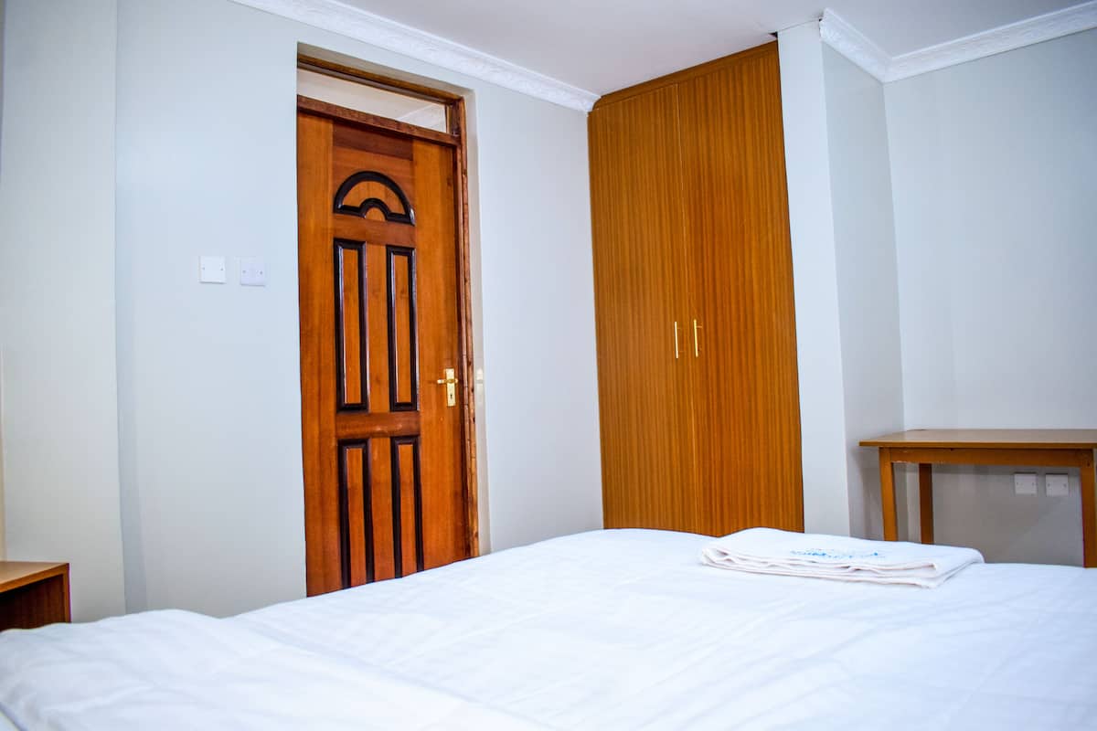 A hotel room with a wooden door, a wardrobe, a bed with white bedding, and a small wooden table.