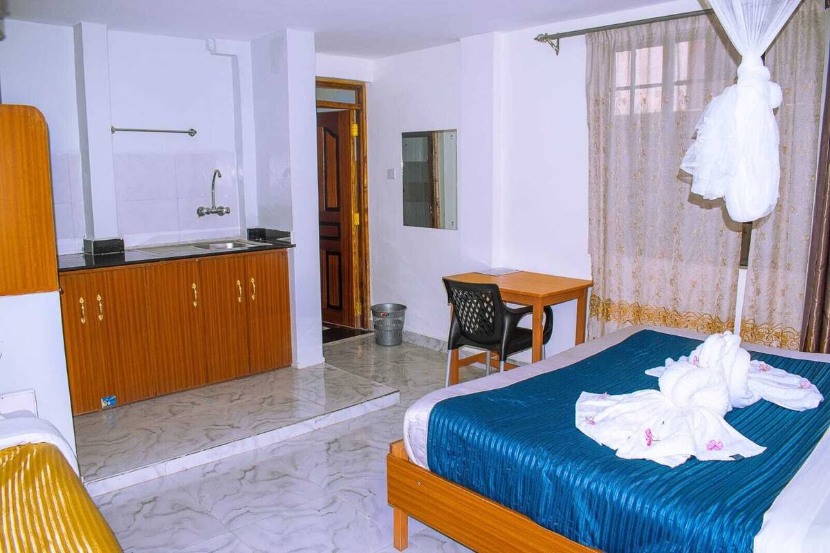 A hotel room with a bed, a small table, a chair, a kitchenette, and a bathroom.