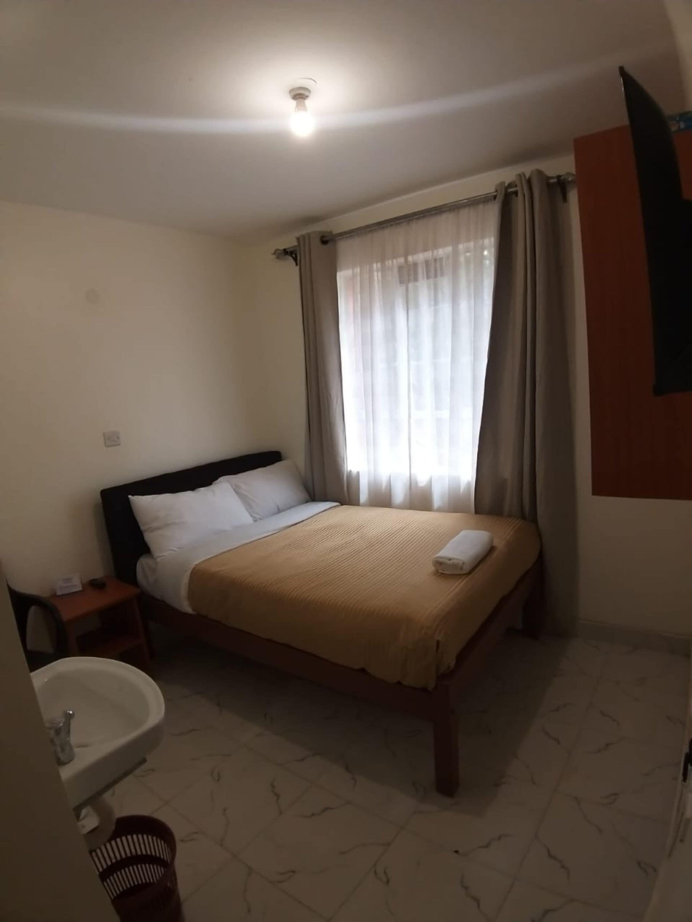 Una habitación de hotel con cama, mesita de noche, baño con lavamanos y espejo, y ventana con cortinas.