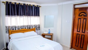 A neatly made bed with white bedding, a wooden headboard, and a bedside table with a bottle and glasses. - Rio Breeze Gardens (Nairobi)