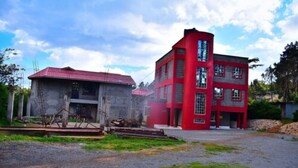 A two-story building with a red facade and a red roof, surrounded by a gravel driveway and greenery. - Rio Breeze Gardens (Nairobi)
