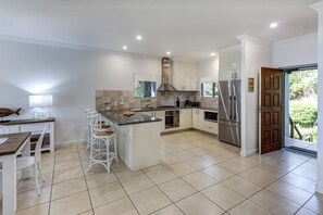 A modern kitchen with a central island, white cabinetry, stainless steel appliances, and a dining area with white chairs. - Blue Water Views 17 (Hamilton Island)