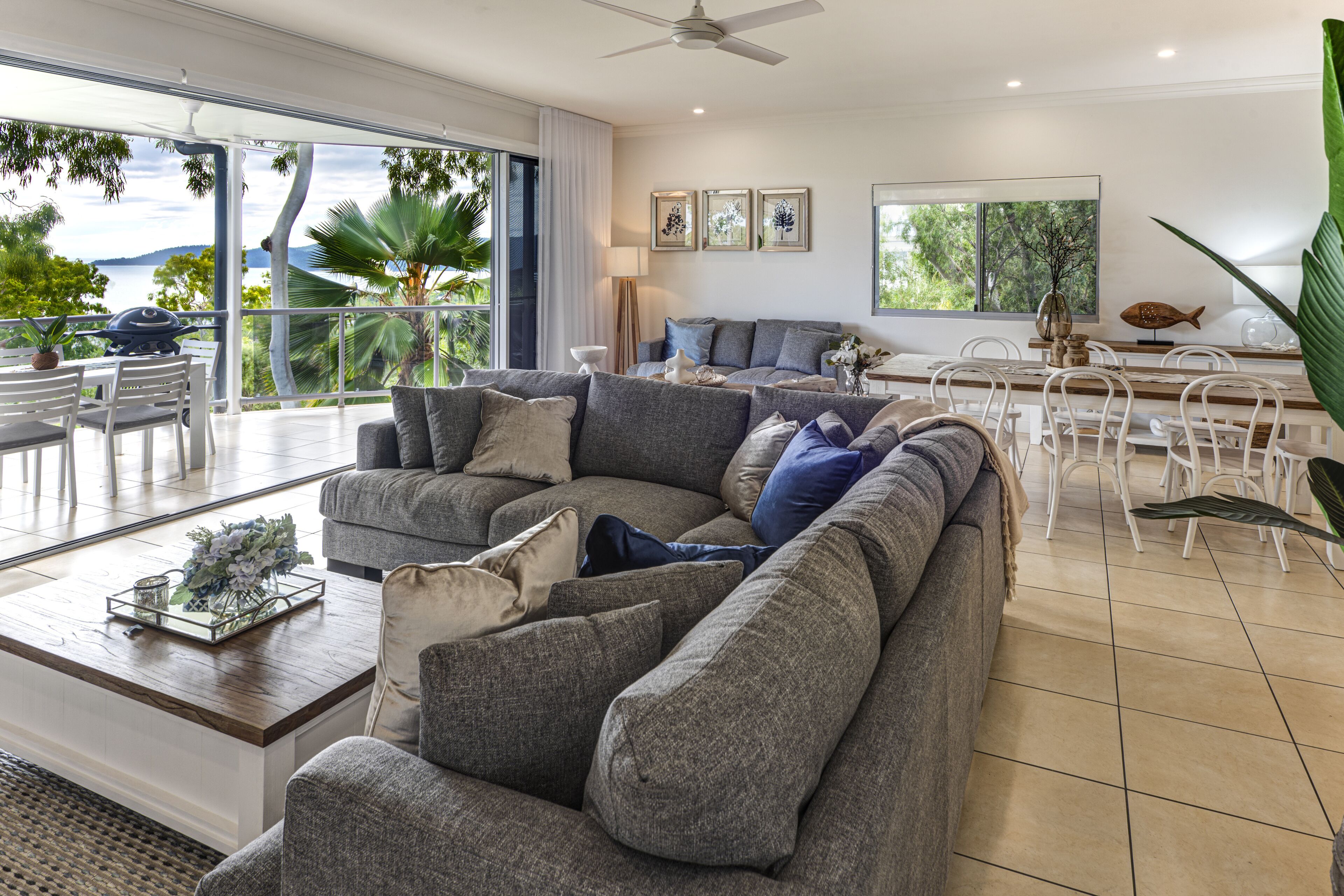 A spacious living area with a large gray sectional sofa, a coffee table with decorative items, and a dining area with white chairs and a wooden table.