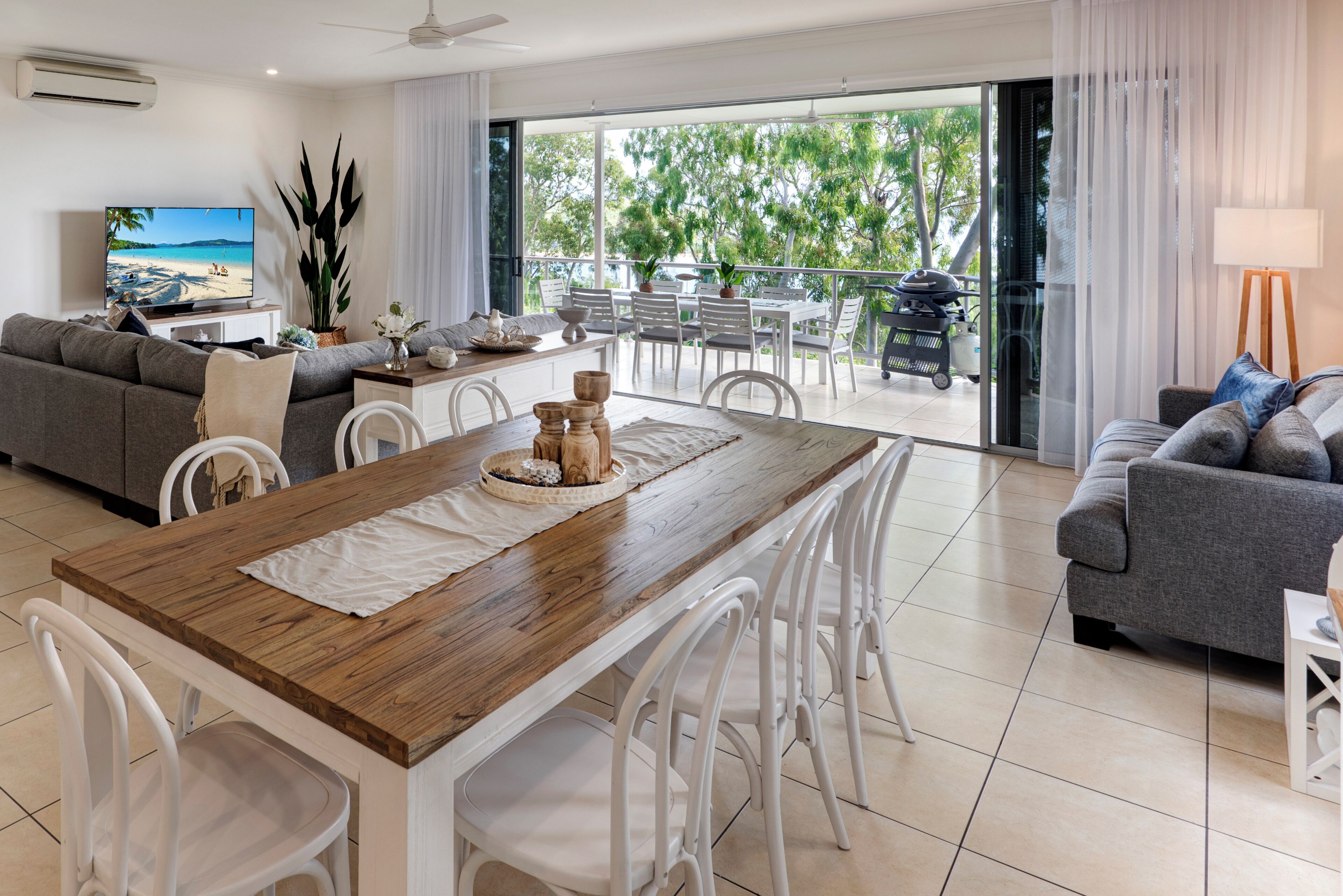 Un'ampia zona giorno con tavolo da pranzo in legno, sedie bianche, divano grigio e un balcone vista alberi e spiaggia.