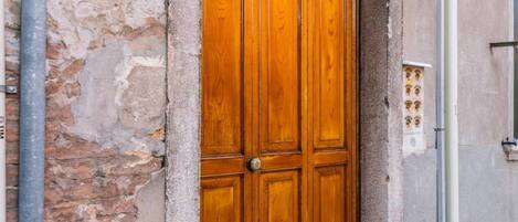 Una puerta doble de madera con un picaporte de metal, empotrada en un muro de piedra con una caja de servicios cercana.