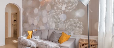 Un salon avec un canapé gris, des coussins jaunes, un lampadaire et un mur orné d’un motif de pissenlit.