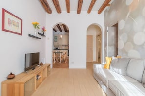 A living room with a TV, wooden console, sofa, and a kitchen in the background.