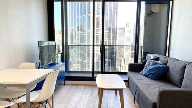 A modern living room with a large window, a grey sofa, a white table, and a TV on a blue cabinet. - Readyset Apartments at Empire (Melbourne)
