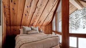 Un dormitorio acogedor con una cama grande, vigas de techo de madera y vistas a un paisaje nevado a través de amplios ventanales.