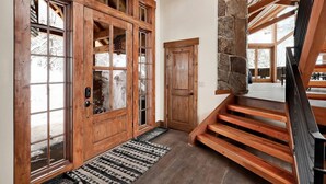 Una escalera de madera que conduce a un segundo piso, un pilar de piedra y una gran puerta de madera con paneles de vidrio.