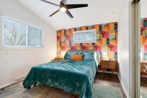 A bedroom with a bed, a ceiling fan, a window with blinds, a colorful wall, and a nightstand with a lamp.