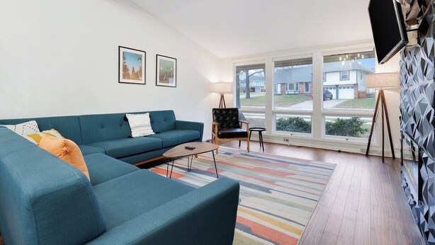 Uma sala de estar moderna com um sofá azul, uma mesinha e um tapete colorido. Há duas imagens emolduradas na parede e uma TV fixada.