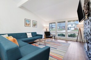Ein modernes Wohnzimmer mit einem blauen Sofa, einem kleinen Tisch und einem farbenfrohen Teppich. An der Wand hängen zwei gerahmte Bilder, und ein Fernseher ist an der Wand befestigt.