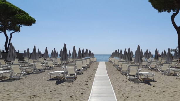 A beach with rows of sun loungers and umbrellas, a wooden walkway leading to the sea, and trees lining the path. - Hibiscus Beach Village - Family apartments within walking distance of the sea (Follonica)
