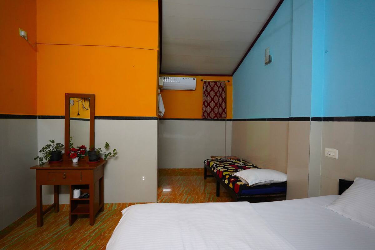 Una habitación de hotel con cama, escritorio con espejo, pequeña mesa con plantas, aire acondicionado de pared y cortina estampada.