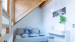Une chambre avec un plafond en pente, un lit avec des oreillers, un petit tabouret, un bureau blanc et une plante.