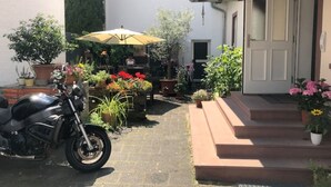 Une moto garée sur un chemin pavé, un patio couvert agrémenté de plantes en pot et une entrée d’immeuble avec des marches.