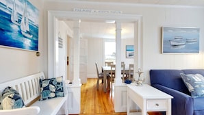Una stanza con una porta bianca con la scritta "Pine Point" e coordinate, un'area pranzo con sedie di legno e un tavolo, un divano blu con cuscini decorati e opere d'arte a tema nautico alle pareti.
