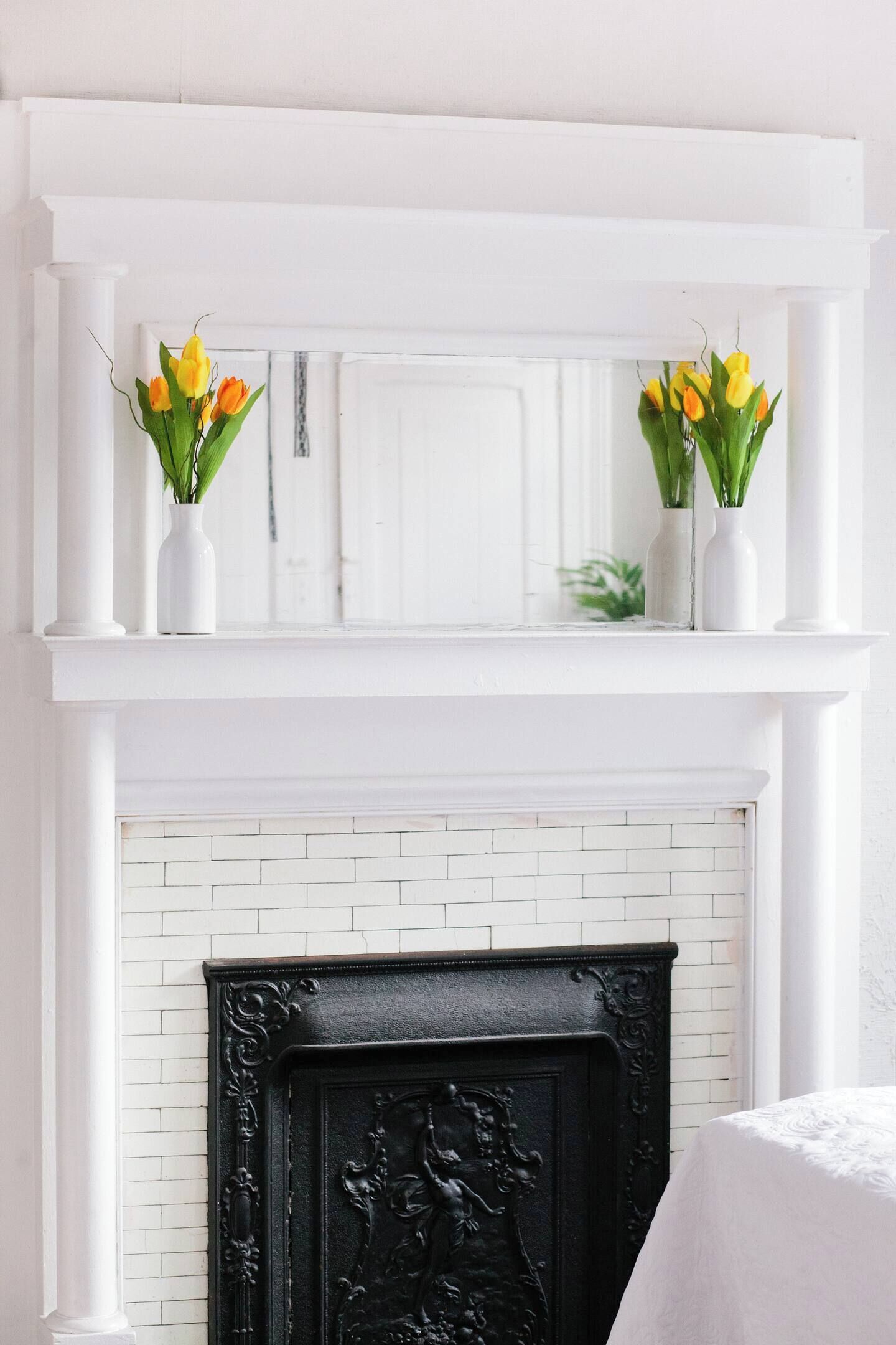 Una chimenea blanca con un marco negro ornamentado, flanqueada por dos estantes blancos con jarrones de tulipanes amarillos.