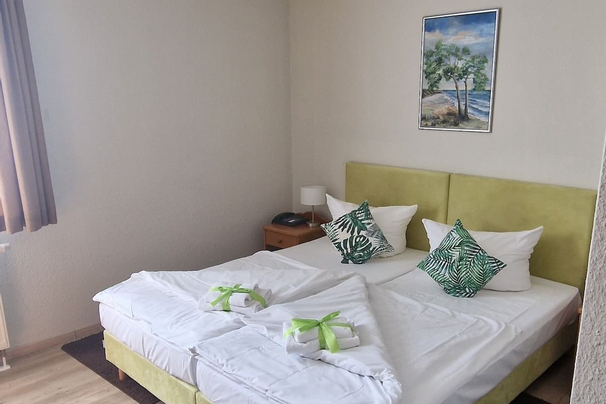 Un lit bien fait, avec du linge de lit blanc et des touches de vert, un tableau représentant un arbre accroché au mur et un plancher en bois.