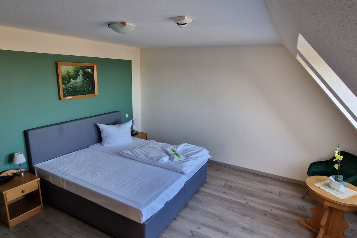 Une chambre à coucher avec un lit, une table de chevet, une petite table sur laquelle se trouve un vase, et un tableau accroché au mur.