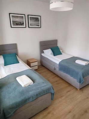 A hotel room with two single beds, each with a grey headboard and teal bedding. There are two framed pictures on the wall above the beds, a small wooden nightstand between the beds, and a modern light fixture above the bed on the right. - Family Apartment in Gdynia | Balcony | Close to Station (Gdynia)